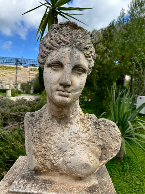 Weathered ancient marble bust statue set in a garden with greenery behind.