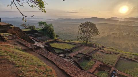 Golden sunrise illuminates Sigiriya rock fortress terraces overlooking misty jungles and distant hills.