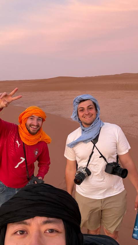 Two young male travelers wearing colorful desert scarves smile for a photo atop reddish sand dunes.