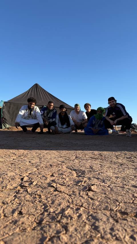 A group of local hosts and travelers kneel together in front of a desert camp tent under blue sky.