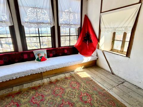 Traditional Albanian interior with long cushioned bench, flag and village view through wooden windows.