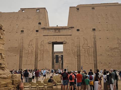 Crowds of visitors entering the massive sandstone pylons of Edfu Temple decorated with carved reliefs of Horus and pharaohs.