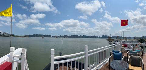 Panoramic deck view of Halong Bay with Vietnamese flag, blue sky, and scattered boats.