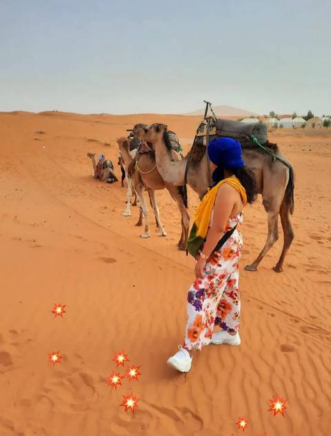 Traveller in colorful dress and blue turban stands beside saddled camels on the orange dunes of the Sahara.