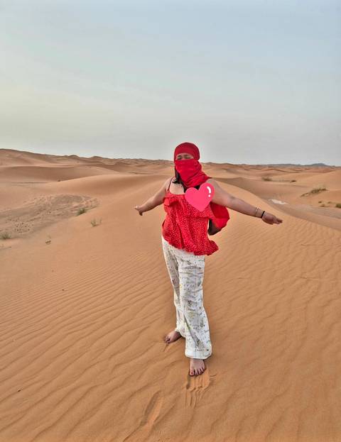 Woman in red scarf spreads her arms joyfully atop endless rolling sand dunes at dusk.