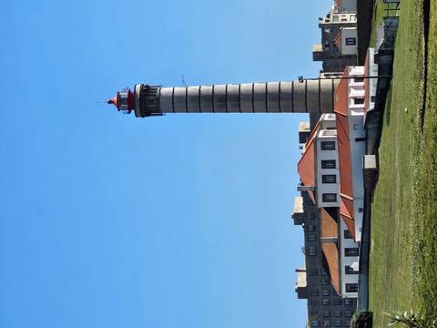 Tall striped lighthouse with red lantern rising above low coastal buildings on a clear day.