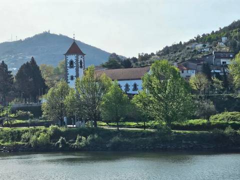 White-walled riverside church framed by leafy trees with hillside village beyond.