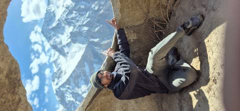 Kneeling hiker points toward towering snow-clad peaks framed by rugged rocks and blue sky.