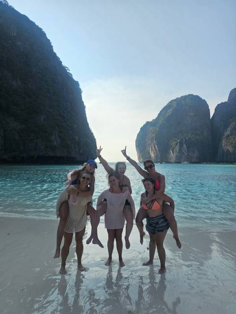 Group of young women posing playfully on a turquoise beach backed by tall limestone cliffs.