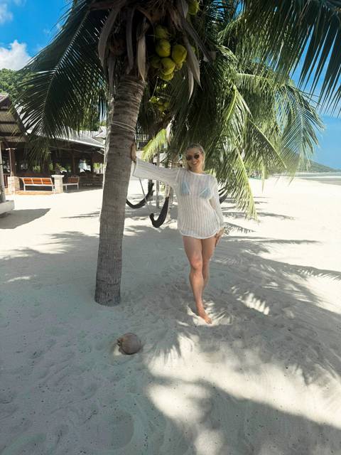 Woman in a mesh cover-up leaning against a palm tree on a white-sand tropical beach.
