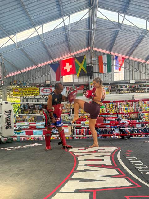 Close-up of Muay Thai sparring as a female fighter knees a pad held by her coach under national flags.