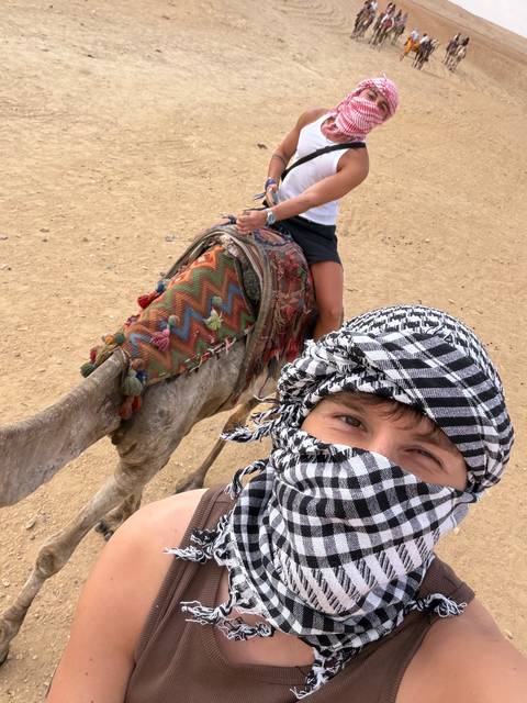 Selfie of a traveller in a checkered keffiyeh riding a camel in the desert.