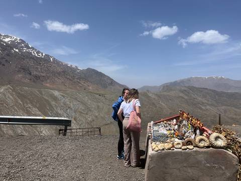 Two travellers browsing a roadside fossil and mineral stall with rugged Atlas Mountains behind