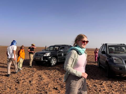 Travellers walk across a dry desert plain near parked SUVs under a clear sky