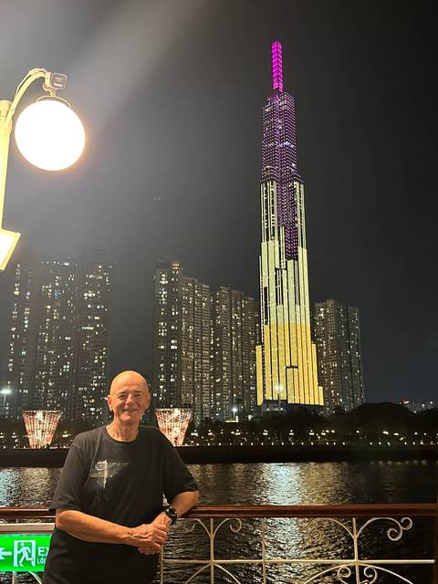 Older traveler posing at night in front of the brightly lit Landmark 81 skyscraper with city high-rises behind.