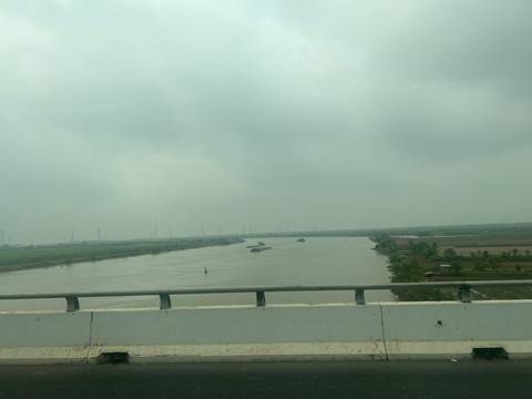Distant barges navigating a broad brown river on a grey cloudy day.