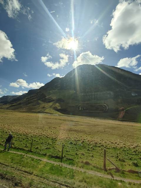 Sun-lit Highland mountain seen through a vehicle window with bright sky and scattered clouds.