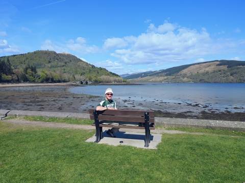 Traveler relaxing on a bench overlooking a tranquil loch and rolling green hills under blue skies.
