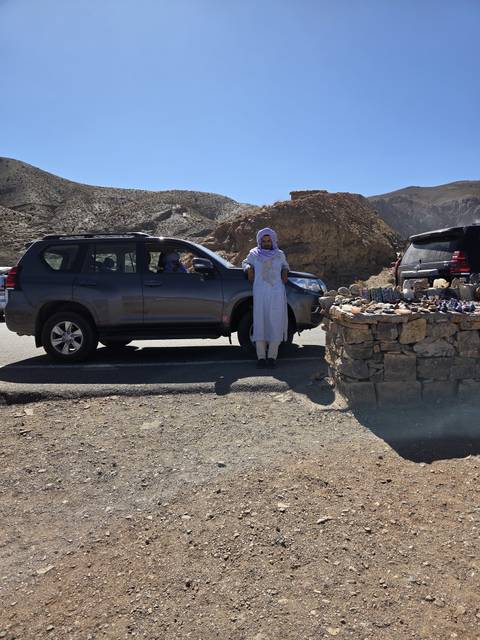 Local man in traditional robe stands beside SUV at roadside stall of stones and trinkets in rugged hills