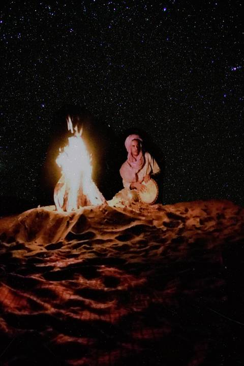 Nighttime desert scene of musician in turban drumming beside a bright campfire under starry sky