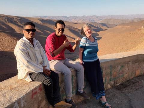 Three friends sitting on a low wall giving thumbs-up with expansive arid mountains and valleys behind them