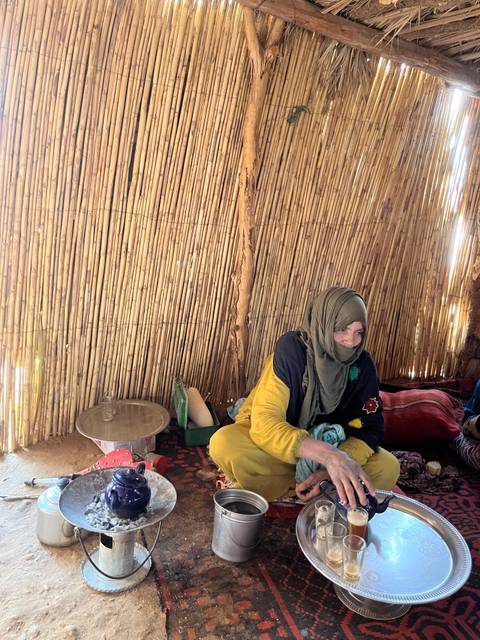 A young woman in traditional dress sits inside a simple reed hut, soft light illuminating the rustic interior.
