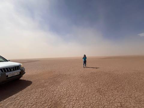 A traveller photographs an immense dry lakebed while a 4x4 vehicle waits at the edge of the frame.