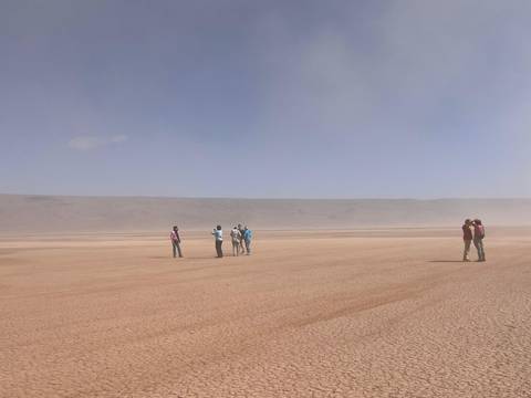 Small groups of travellers stand on a vast, dusty clay pan under a hazy blue sky.