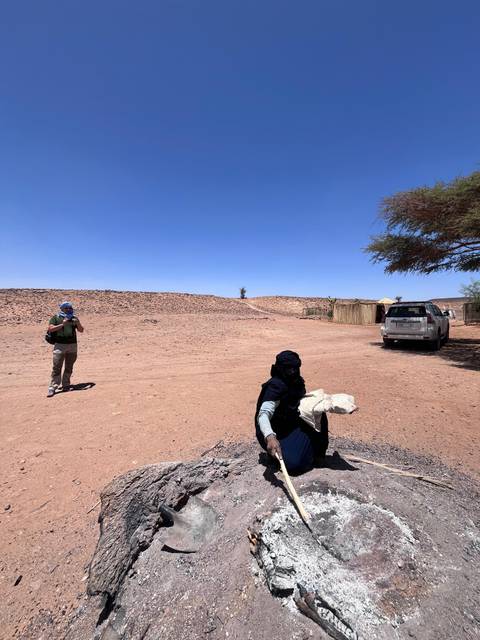 Traveller wearing a scarf photographs a desert settlement while a local man in turban kneels in foreground.