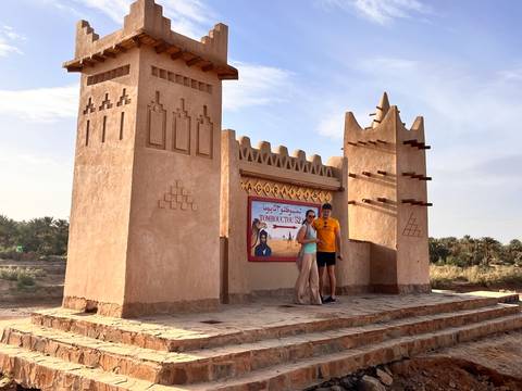 Couple posing by an adobe-style desert gate decorated with Arabic script and palm groves beyond.