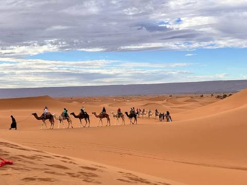 Long camel caravan led by a guide traverses golden Sahara dunes beneath a patchy sky.