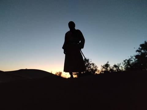 Silhouette of a person in traditional robe holding a stick atop a dune at dawn.