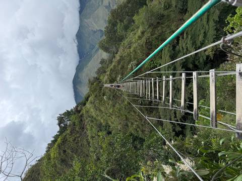Narrow hanging bridge made of wooden planks and cables stretches across a deep green gorge toward distant hills.