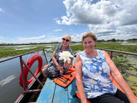 Two women on a small wooden boat in a lily-covered lake, one holding a white lotus bloom.