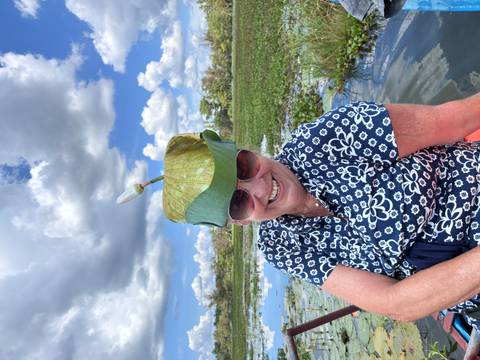 Woman laughing while wearing a whimsical lotus-leaf hat, with wetlands and blue sky behind.