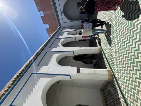 Visitors walk through an arched courtyard with patterned green-white tiles under a clear Marrakech sky.