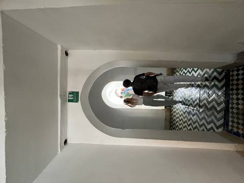 Tourists walk down a narrow arched corridor with patterned floor tiles toward daylight.
