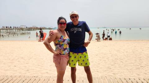 Smiling man and woman pose on a sandy beach beside shallow turquoise water with a pier and bathers in the background.