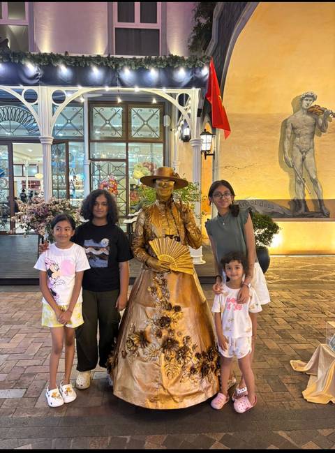 Children pose smiling beside a golden living-statue street performer holding a fan in a festive pedestrian area.