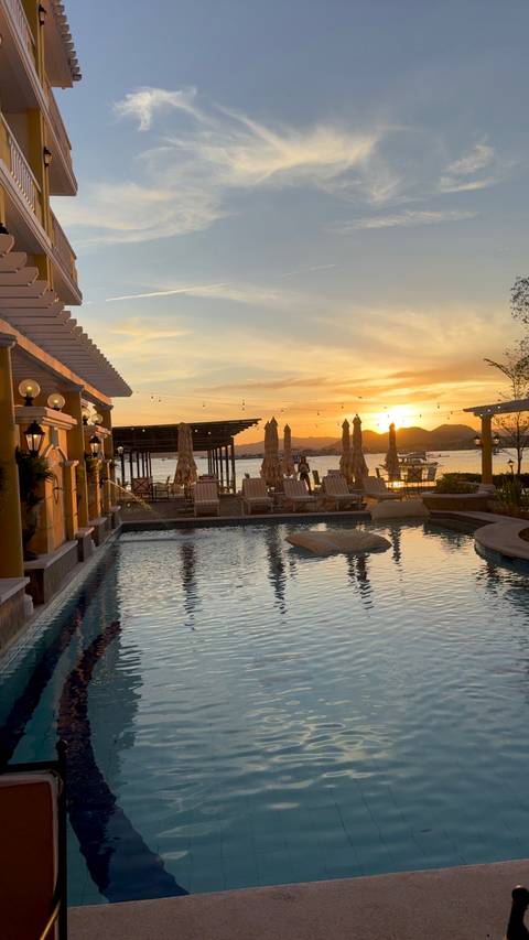 Resort pool bordered by deckchairs and sun umbrellas overlooking a bay and mountain silhouettes at golden sunset.