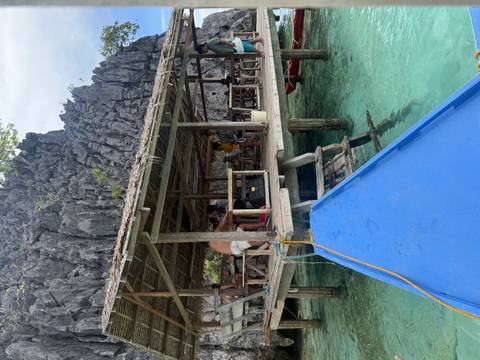 Rustic waterside hut on stilts beside dramatic limestone cliffs, viewed from the bow of a blue outrigger boat.