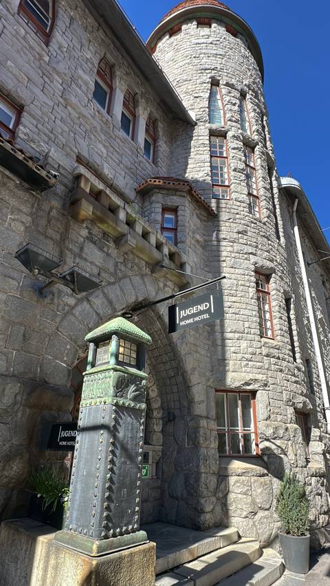 Black sign for Jugend Home Hotel hangs from an arched stone building with small windows.
