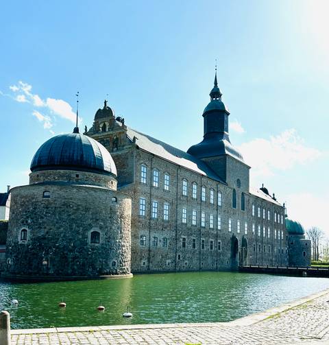 Imposing stone castle with rounded towers and tall spire set against a bright sky.