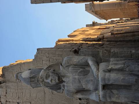 Huge seated pharaoh statue outside temple walls warmly lit by early morning sun with carved hieroglyphs in background.