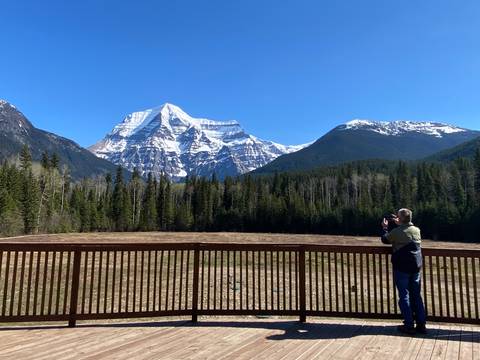 Photographer on a viewing deck capturing the snow-covered peak of Mount Robson rising above dense forest