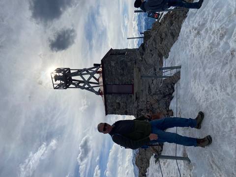 Hiker standing on a snowy summit beside an old stone weather station silhouetted against the sun