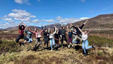 Joyful group of travellers captured mid-jump in an open Highland landscape under blue skies.