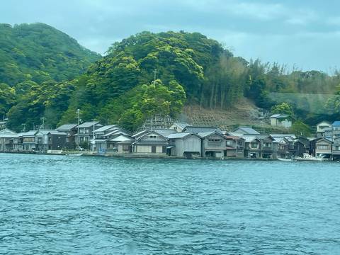 Traditional wooden boathouses lining a quiet coastal village backed by dense green hills.