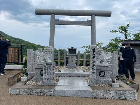 Granite torii gate and shrine monument atop a hill with travellers taking photos and hazy views beyond.