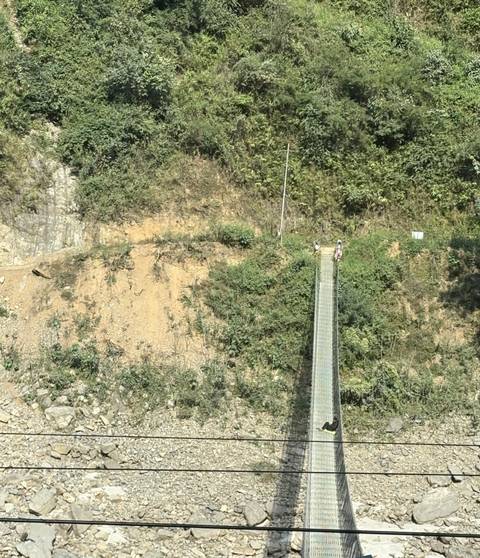Two trekkers crossing a long narrow suspension bridge over a deep forested ravine.